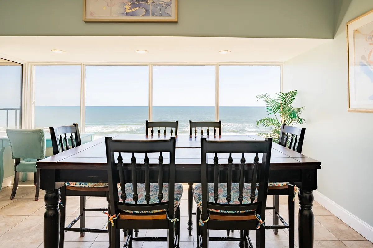 A bright dining area features a table set beside expansive windows framing a captivating ocean view. Lush greenery and vibrant wall art add a sense of comfort and warmth, while the natural light highlights the room’s joyful, tranquil ambiance.