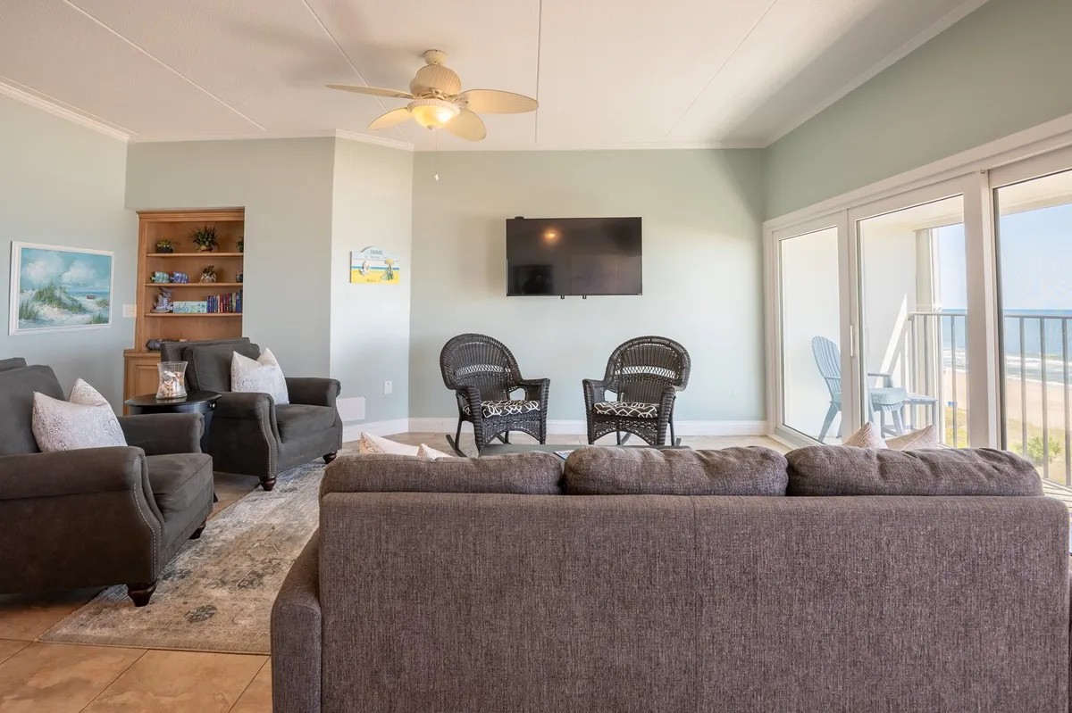 This image showcases a comfortable living area with a grey sofa, wicker chairs, a flat-screen TV, and expansive windows that bring in natural light and offer a clear ocean view. A bookcase with books and inviting decor suggest a space perfect for relaxation and gathering.