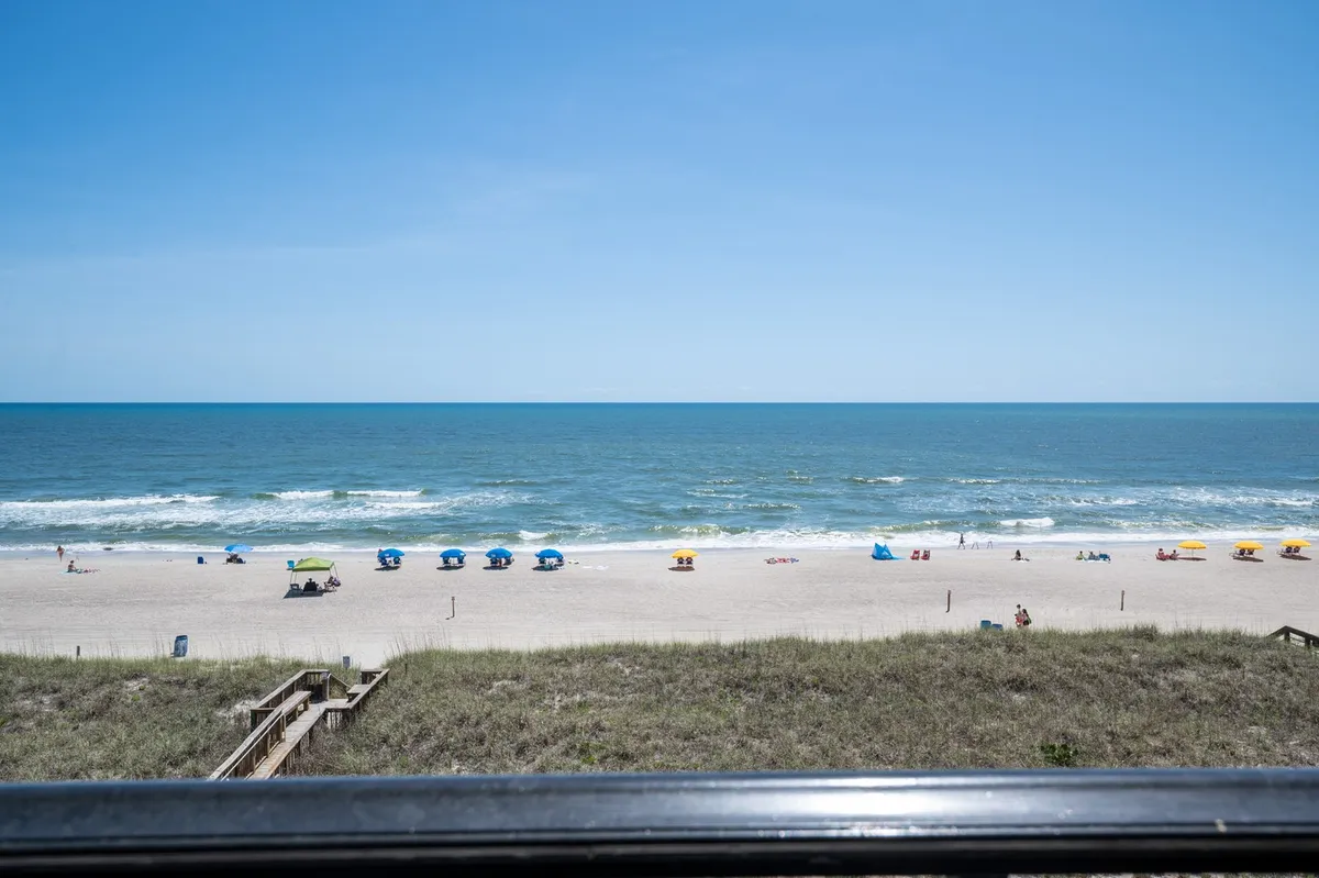 A wide, sunlit view of the sandy beach and ocean, with colorful beach chairs arranged facing the waves under a clear sky. The scene conveys activity options such as sunbathing, swimming, and family gatherings by the sea.