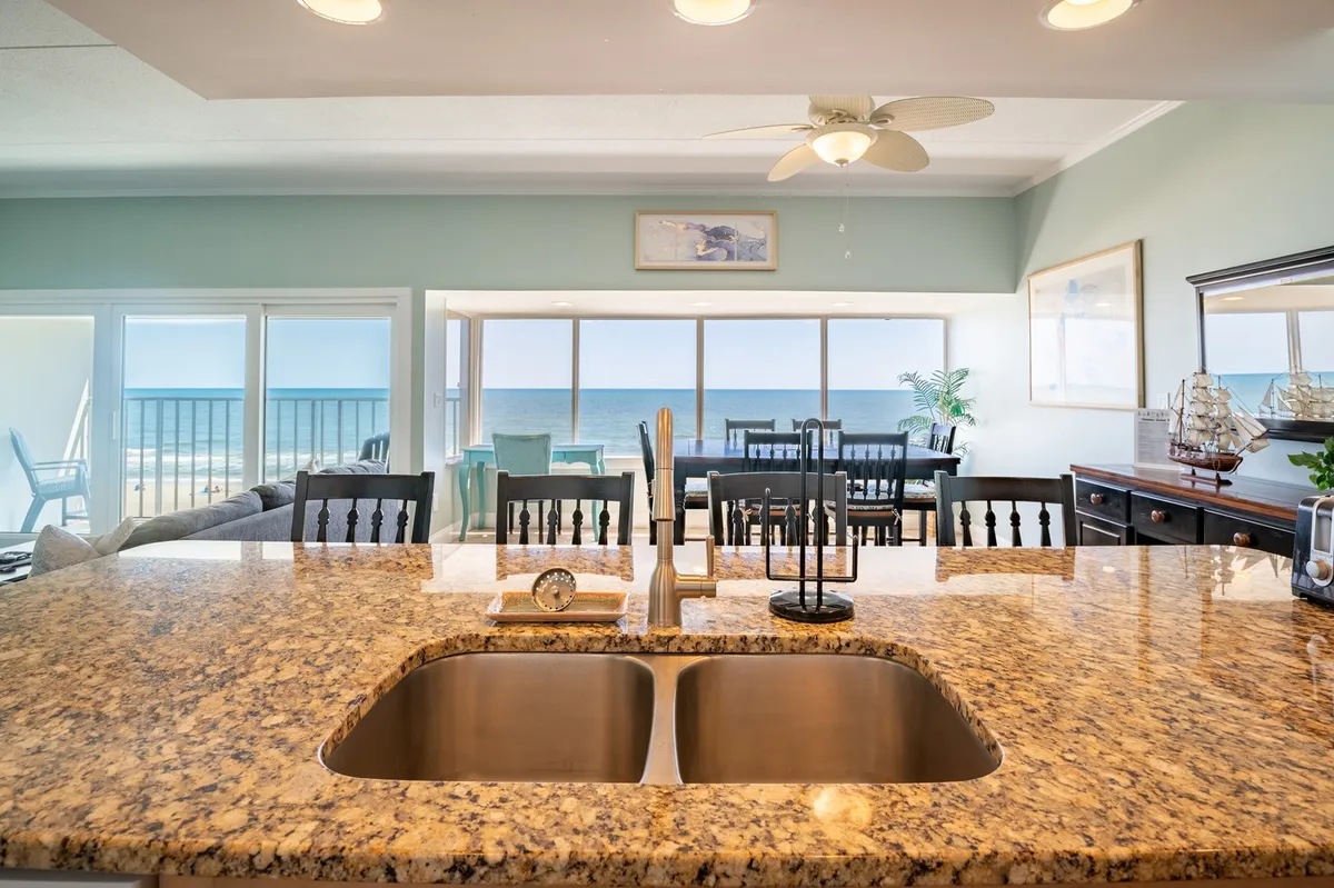 This interior view shows a granite-topped kitchen counter overlooking both the kitchen sink and a dining area with ocean views through the windows. The space combines food preparation and socializing, with natural light and scenery enhancing the atmosphere.