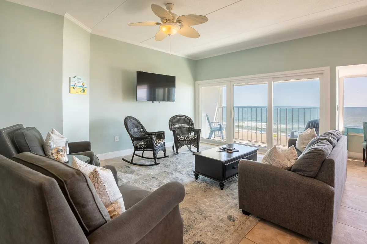 The image showcases a bright, airy living room with large windows revealing breathtaking ocean views. Plush, comfortable seating surrounds a stylish coffee table, inviting guests to relax and socialize while enjoying the coastal scenery. The space is filled with natural light and features a ceiling fan, making it the perfect spot to unwind or gather with loved ones.