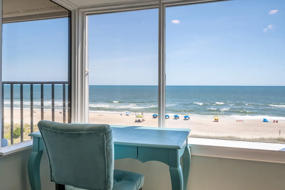 A sweeping ocean view dominates the frame, with the shoreline and gentle waves visible in the distance. A desk sits near the window, providing a tranquil spot to write, relax, or take in the sunrise. The composition emphasizes the connection to the outdoors and the inviting coastal atmosphere.