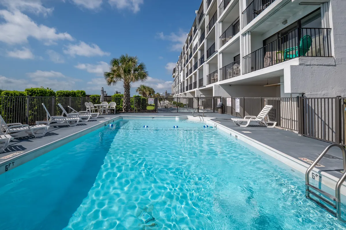 A sparkling swimming pool sits oceanfront, framed by sun loungers and palm trees, with the condo building rising in the background. The area invites relaxation and social gatherings, with ample seating for guests and direct views of the ocean beyond.