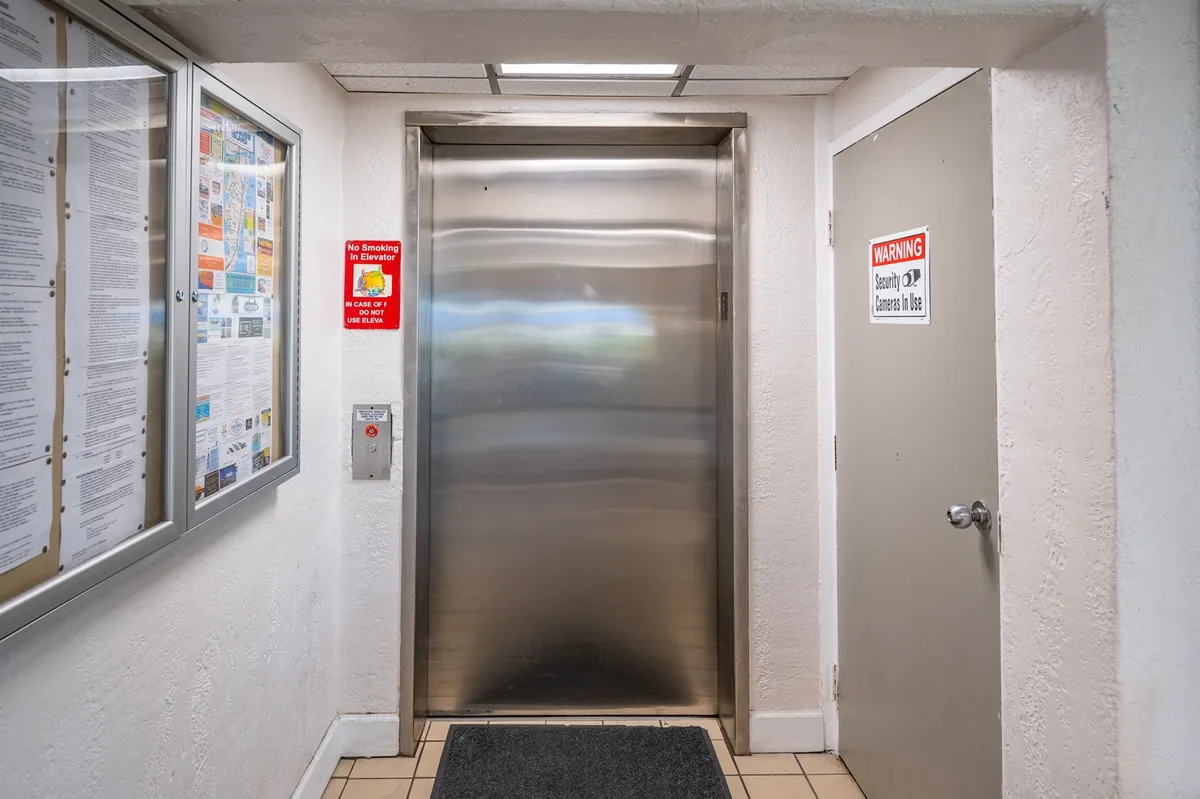 A neutral-toned elevator door is set beside an information board and a warning sign, in a well-lit building common area. The setup is practical, emphasizing ease of movement between floors while nodding to operational notes about the elevator.