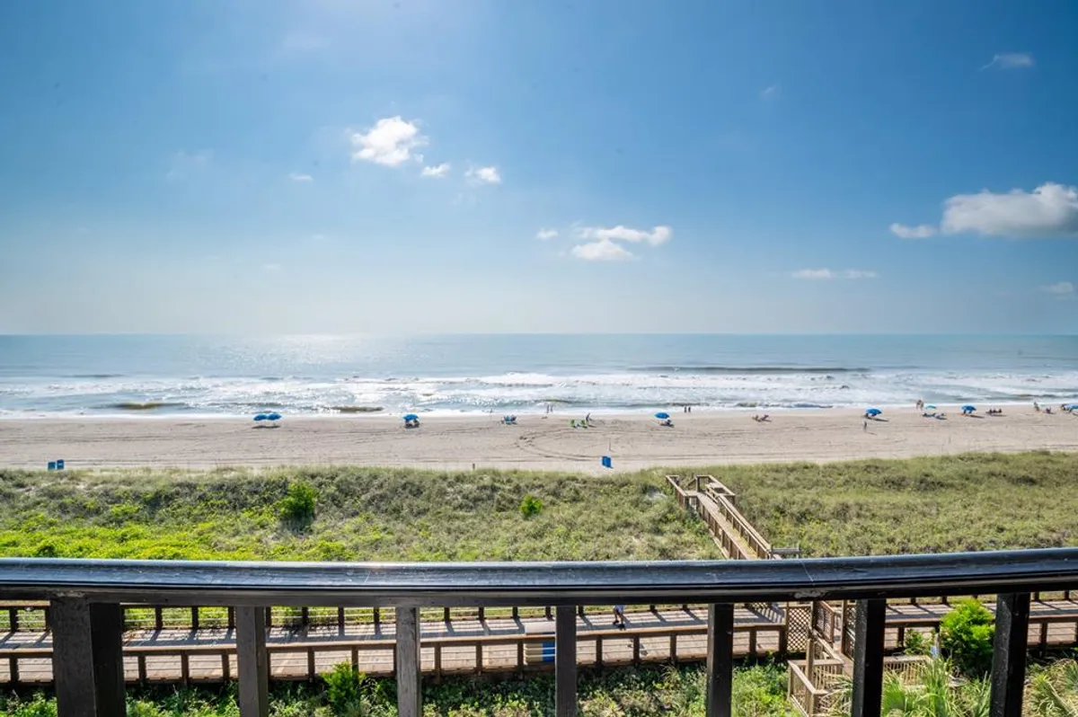 A picturesque scene featuring the ocean, sandy beach, and a wooden boardwalk leading directly to the shore. Umbrellas dot the sand, and the inviting water stretches out under a sunny sky, showcasing the ease of beach access from the community.