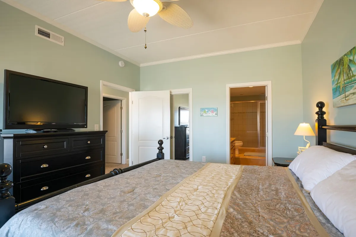 A serene primary bedroom featuring a king-sized bed with crisp linens, a nightstand with a lamp, a television on the opposite wall, and thoughtfully chosen artwork. Natural light fills the room, creating a restful, inviting ambiance.
