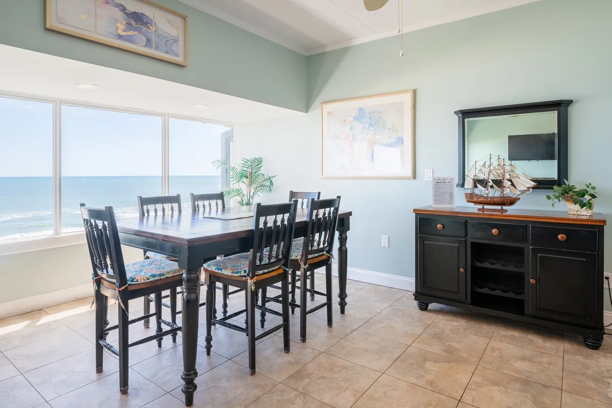 The dining area features a large table set near windows that frame serene ocean views. Decorative elements like a black cabinet with a ship model and wall art give a touch of coastal charm, while the table invites family meals and games.