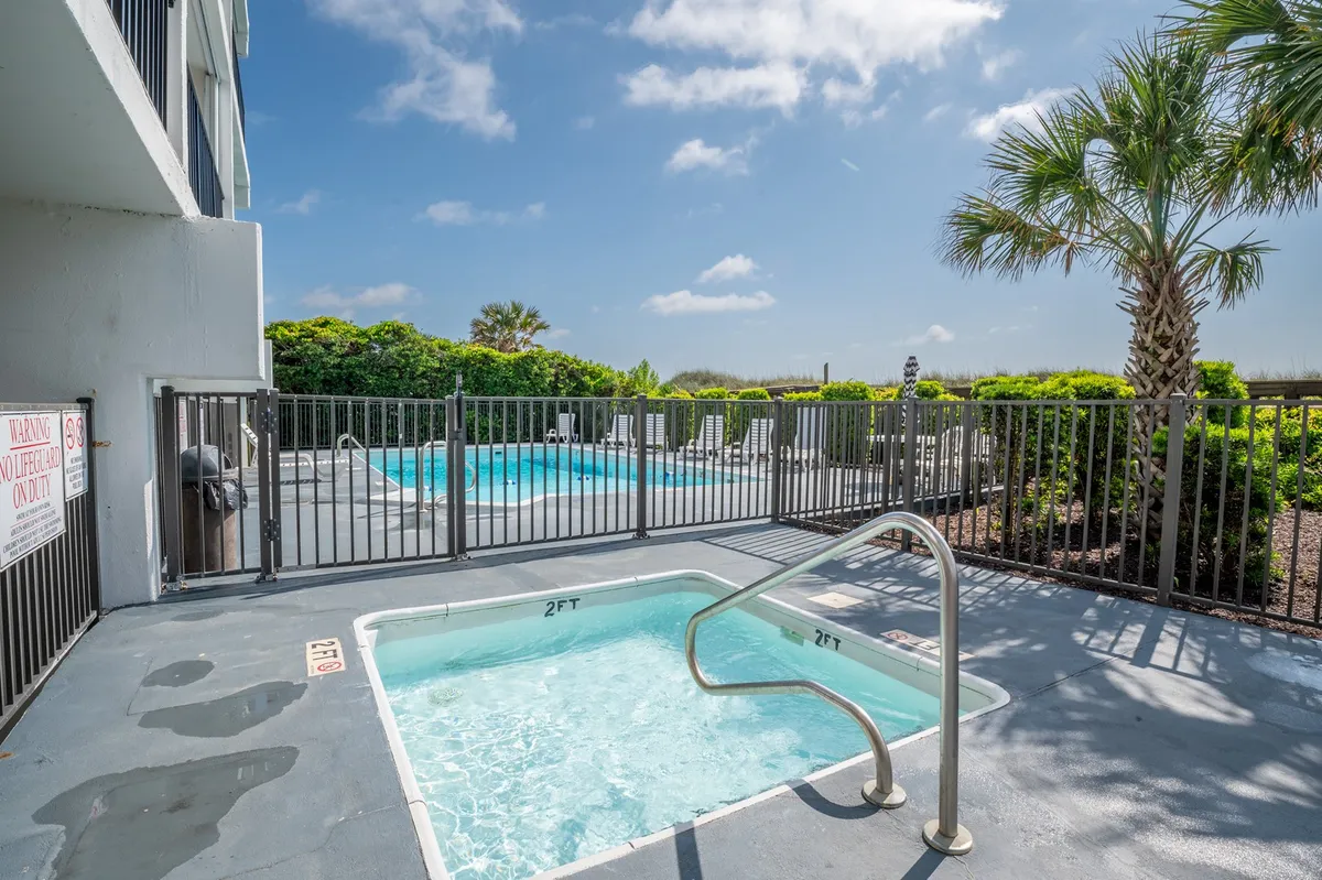An inviting exterior pool area with a sparkling swimming pool, hot tub, and surrounding palm trees. Lounge chairs line the deck, creating a relaxing and safe spot for families to unwind after a day at the beach.