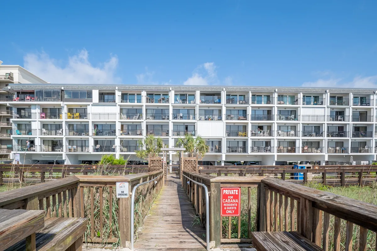 A wooden boardwalk leads toward the condo building, flanked by palm trees and balconies adorned with colorful chairs. The walkway suggests direct beach access, inviting residents and guests to easily transition from the property to the sand.