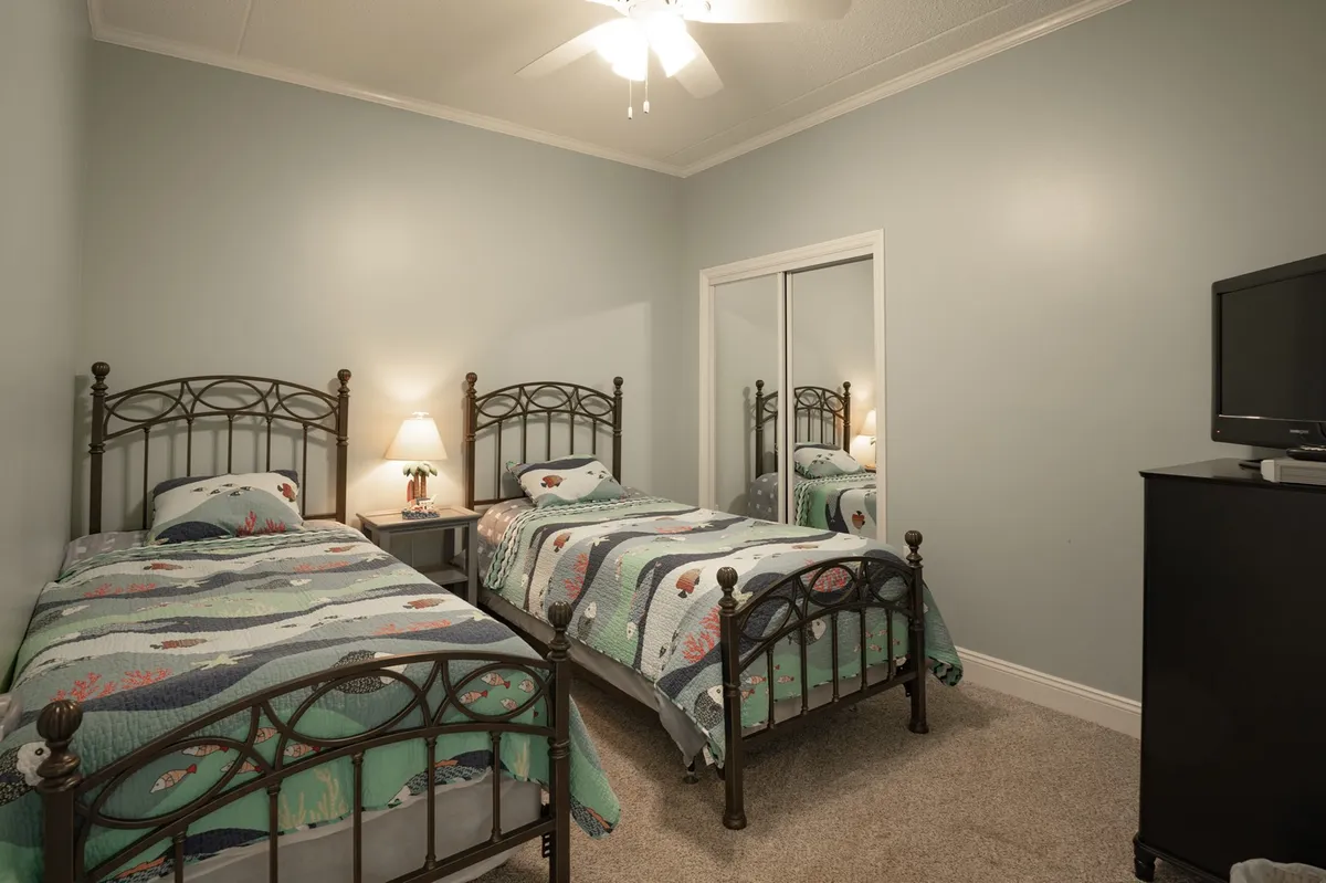 A guest bedroom featuring two twin beds with colorful quilts, a nightstand with a lamp, mirrored closet doors, and a television. The room looks bright and cozy, providing an ideal retreat for guests or children.